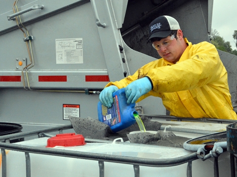 a man in a yellow jacket pouring motor oil into a large disposal container 