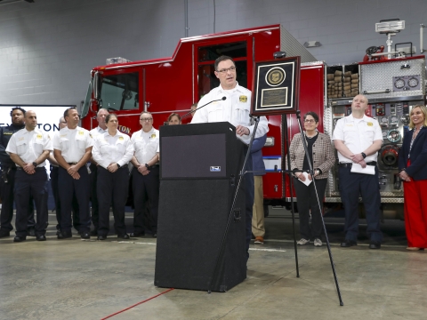 Chief Jason Wells speaks at a lectern with members of the fire department behind him. 
