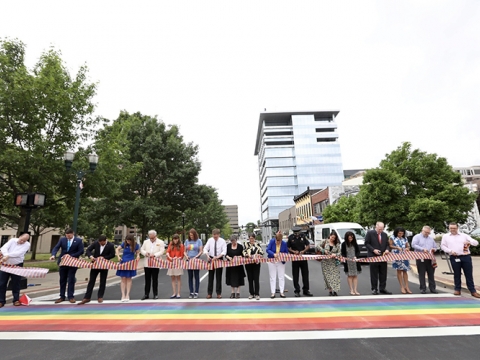 Mayor Linda Gorton, with about 20 community leaders, stand at a rainbow-colored crosswalk at an intersection in downtown Lexington while cutting a large ribbon.