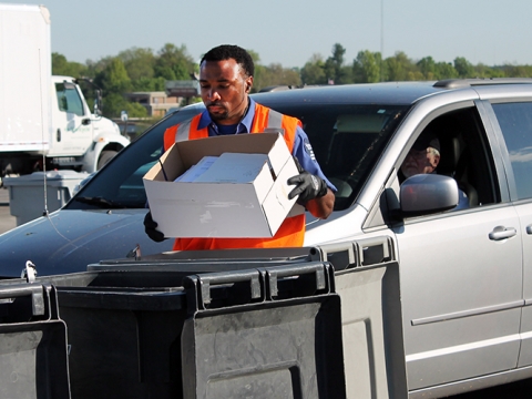 man wearing a safety vest outside emptying box of white paper into a bin with a line of cars behind him