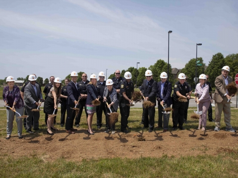 Mayor Linda Gorton, members of the Urban County Council and police officers stand side by side shoveling dirt in an open field on a sunny summer day