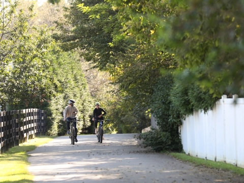 2 cyclists riding toward the camera on the paved Brighton Trail, surrounded by trees with a wooden fence on one side, on a sunny day
