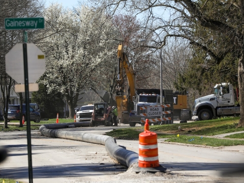 a photo of an intersection with the Greentree street sign visible; there are orange barrels and a large pipe in the street with construction equipment in the background